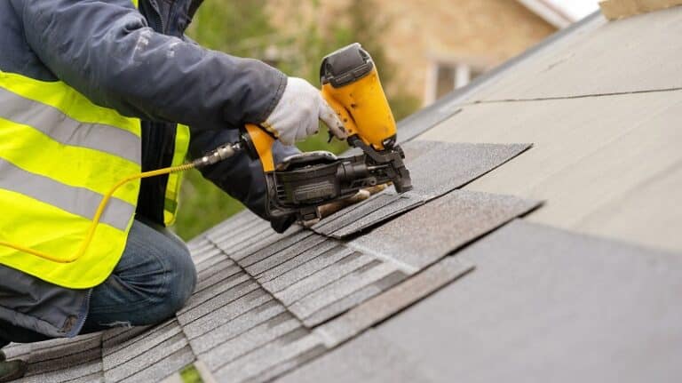 Worker in a reflective vest using a nail gun on a shingled roof.