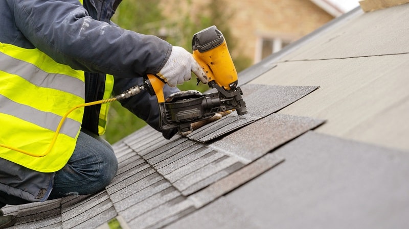 Worker in a reflective vest using a nail gun on a shingled roof