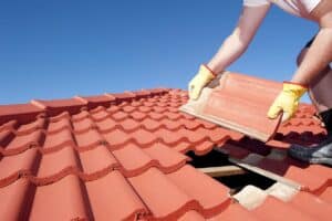 Person in gloves fixing a red tiled roof under a clear blue sky.