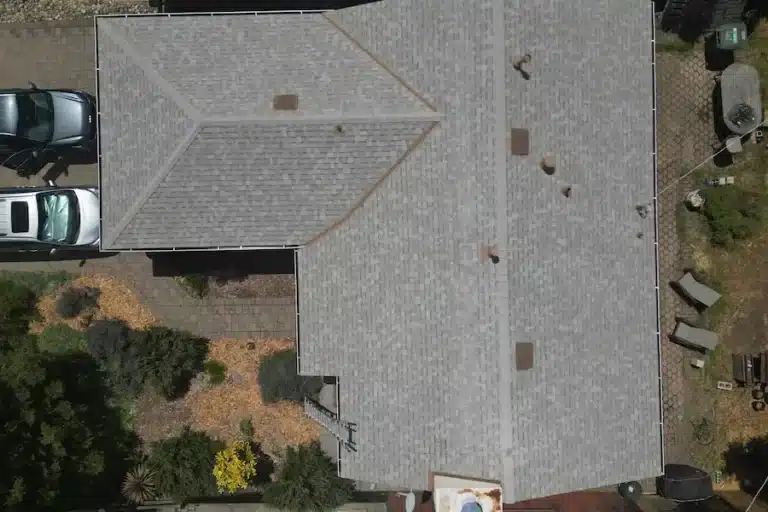 Aerial view of gray shingled roof with skylights, landscaping, patio, and parked cars around