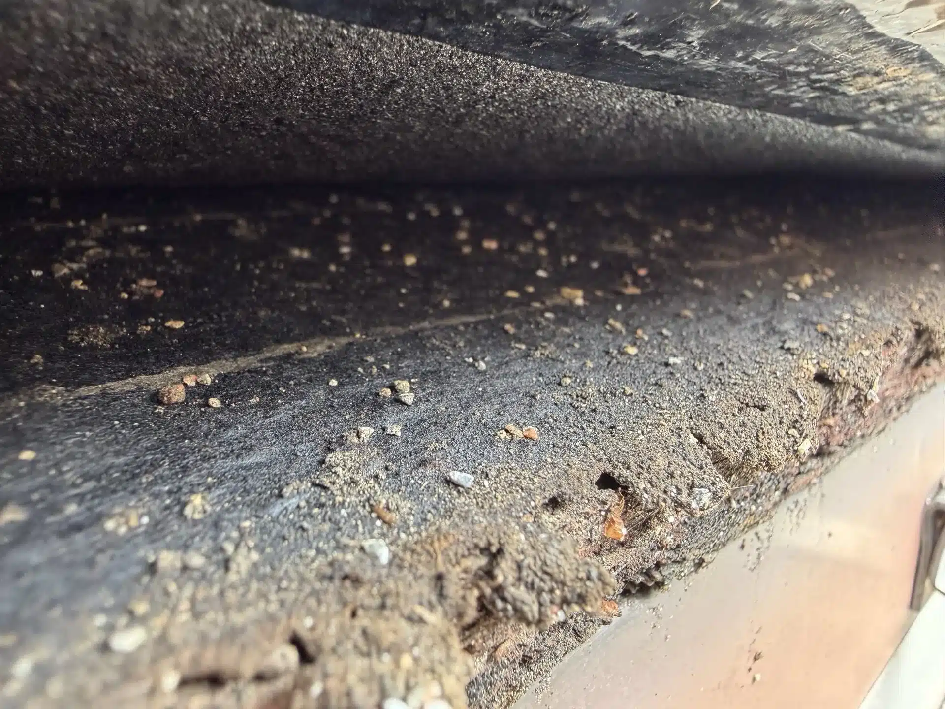 Close-up of an aging asphalt shingle edge with heavy granule loss and a granule-filled gutter