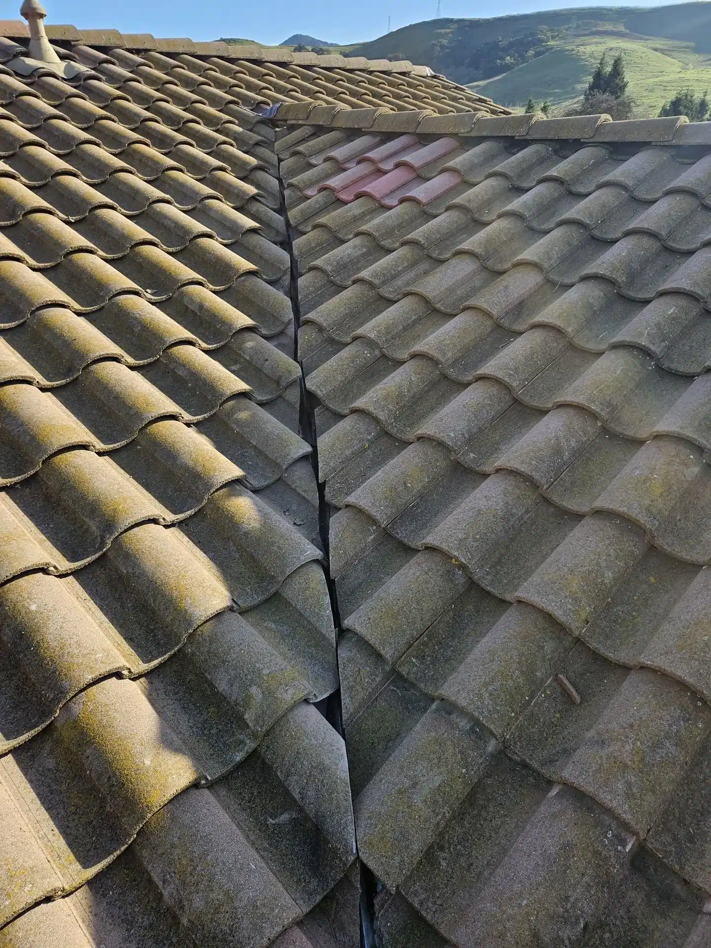Two moss-covered concrete tile planes meeting at an open valley on a Morgan Hill roof, with mismatched replacement tiles near the ridge